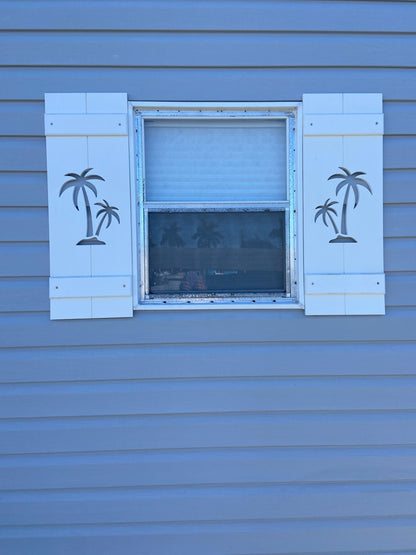 Window with decorative shutters featuring palm tree designs on a blue siding.